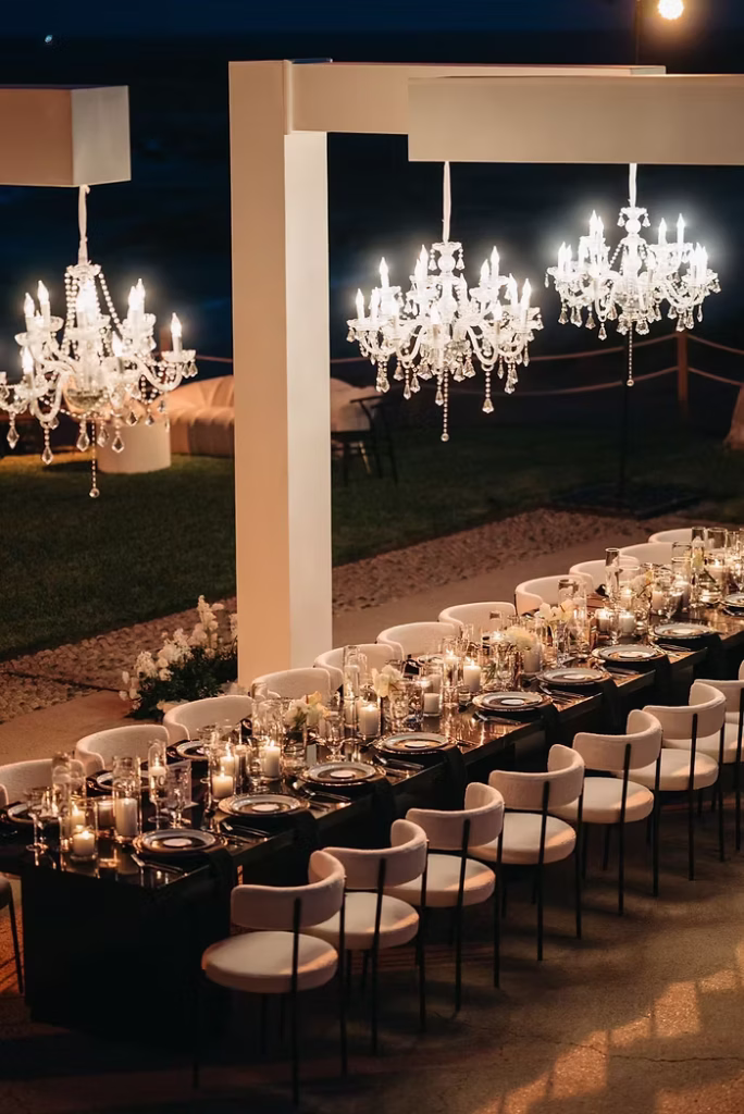 Elegant wedding reception table setting with chandeliers and candles at night.